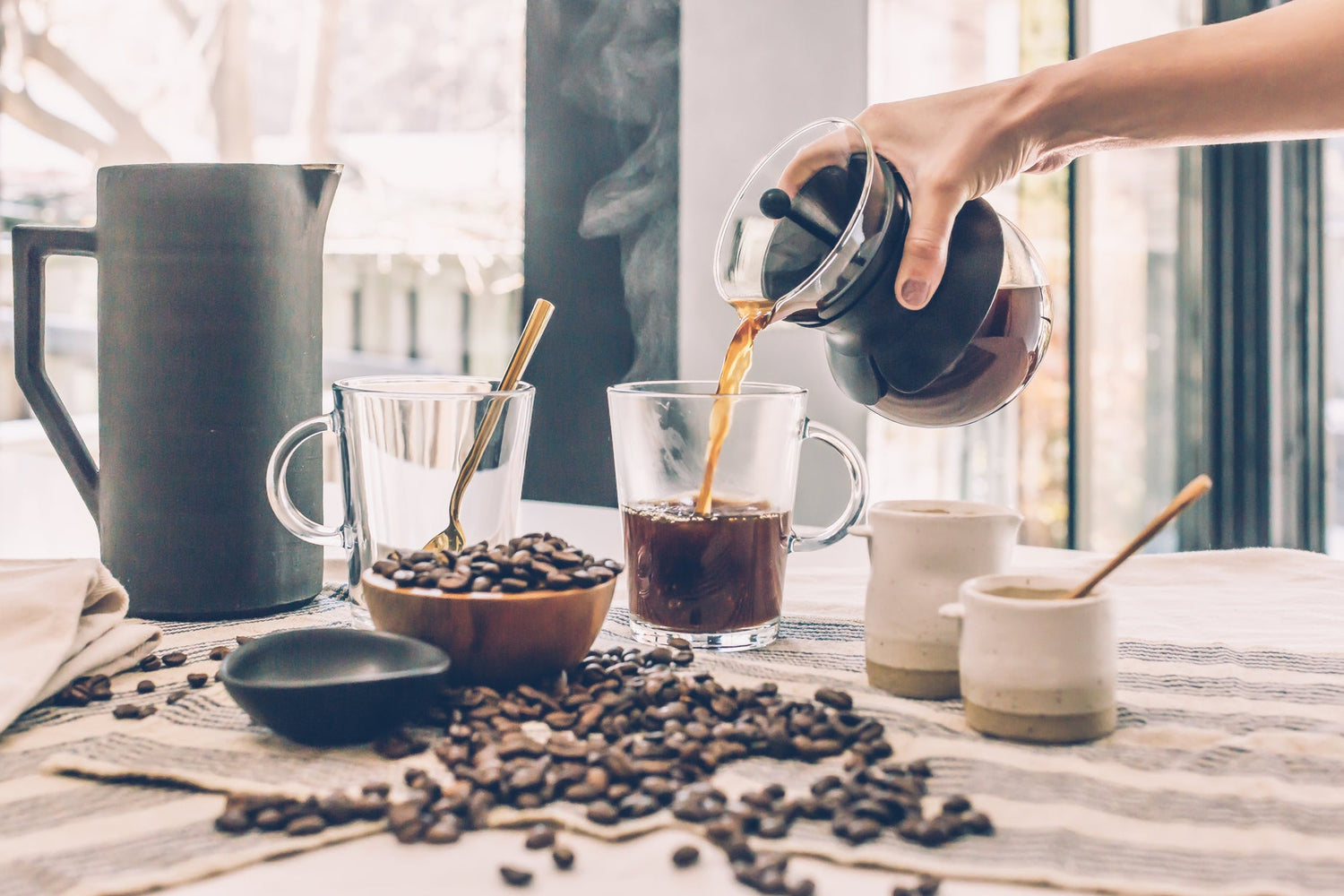 Hand pouring coffee from a French press into a glass mug on a table with coffee beans, a ceramic pitcher, and a bowl of coffee beans in the background