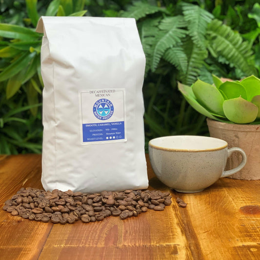 White bag of Leaf and Vine decaffeinated Mexican coffee by Mountain Water Process on a wooden table, surrounded by coffee beans, with a ceramic cup and a potted succulent in the background