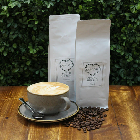 Two 227g white bags of Leaf & Vine Machu Supreme coffee, one ground and one whole beans, on a wooden table with a latte in a gray cup and saucer, surrounded by coffee beans, with a green leafy background