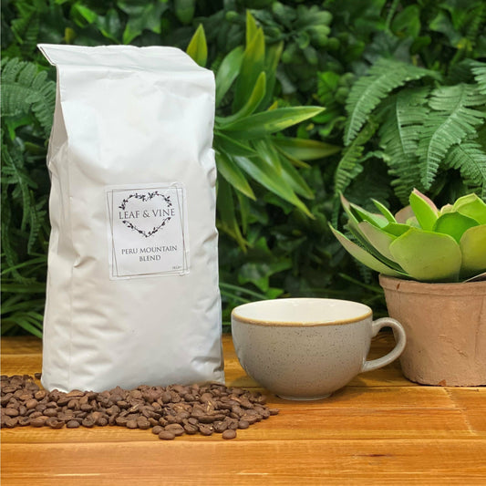 White bag of Leaf & Vine Peru Mountain Blend coffee on a wooden table surrounded by coffee beans, with a ceramic cup and a potted succulent plant in the background