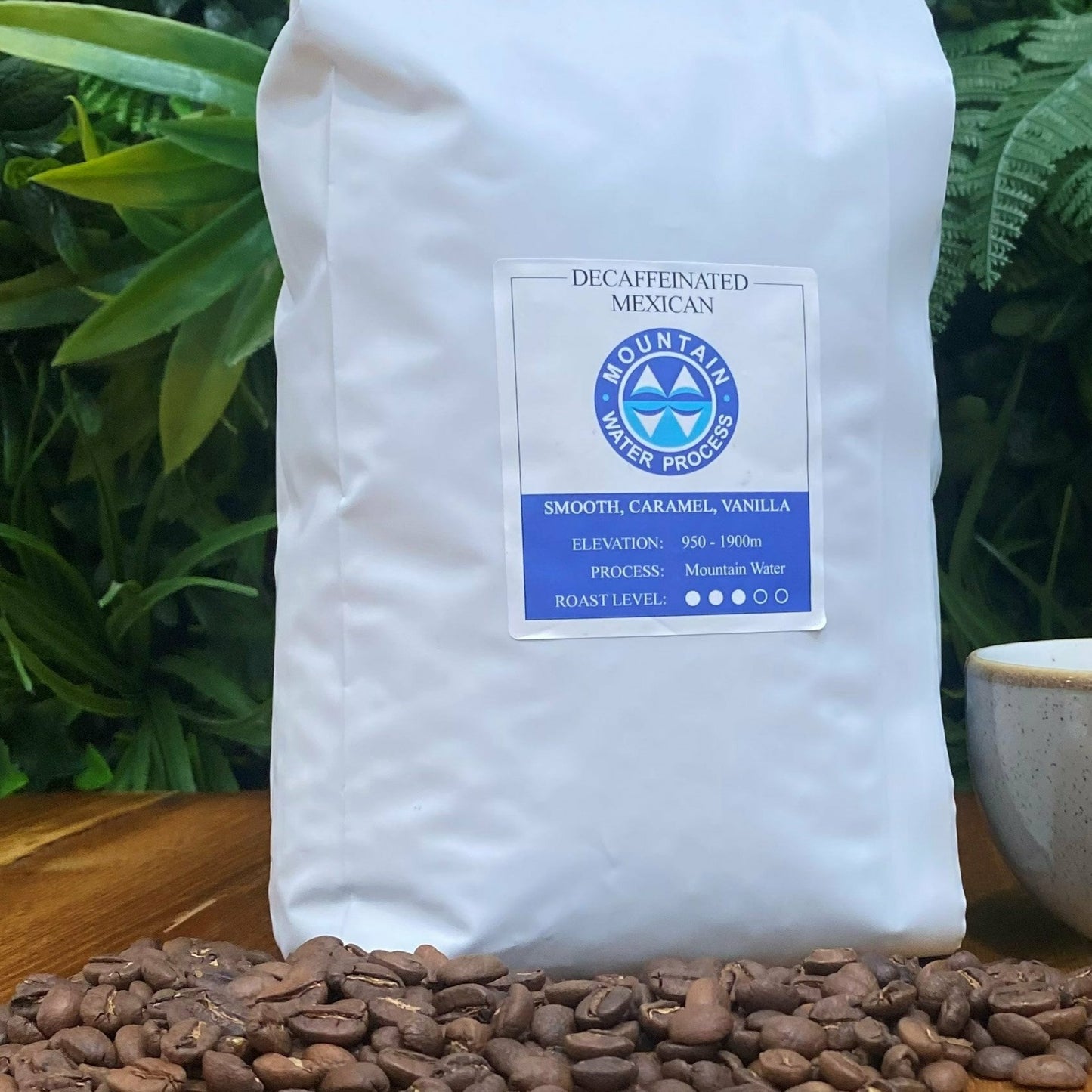 White bag of Leaf and Vine decaffeinated Mexican coffee by Mountain Water Process on a wooden table, surrounded by coffee beans, with a ceramic cup and a potted succulent in the background